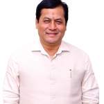 Sarbananda Sonowal, former Chief Minister of Assam, smiling in formal attire, with the Indian flag in the background, symbolizing his leadership and service.
