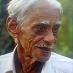 Portrait of Hiren Bhattacharyya, an elderly Assamese poet with white hair, wearing a white shirt, and smiling warmly against a natural green background.
