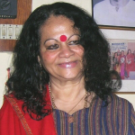 A photograph of Indira Goswami, the renowned Assamese writer and activist, wearing a red bindi and a traditional outfit, smiling warmly.