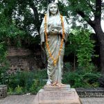 A statue of Sati Joymoti adorned with a garland, standing in a garden with lush greenery and a historical brick wall in the background.