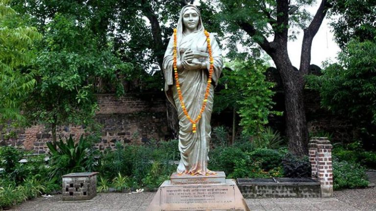A statue of Sati Joymoti adorned with a garland, standing in a garden with lush greenery and a historical brick wall in the background.
