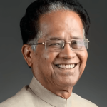 A photograph of Tarun Gogoi, former Chief Minister of Assam, smiling in a formal suit and glasses, taken during a public event.