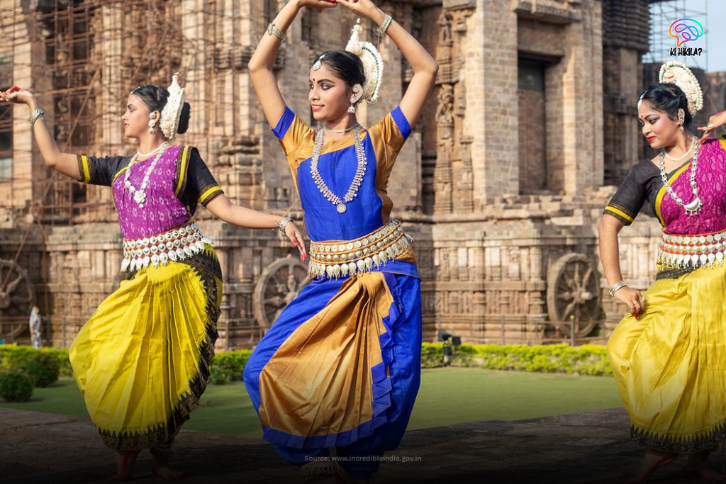 Three female dancers in vibrant, traditional costumes performing a classical Indian dance in front of an ancient, carved stone temple.