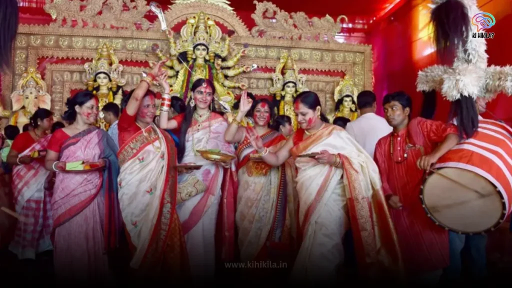 A group of women in traditional sarees celebrating with sindoor and a man playing a dhaak (drum) during Durga Puja 2025.