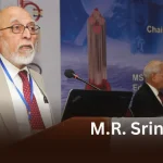 A photo of M.R. Srinivasan, a distinguished scientist, speaking at a podium with a conference banner in the background.