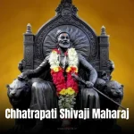 A dark stone statue of Chhatrapati Shivaji Maharaj seated on a throne adorned with flower garlands, set against a golden background.