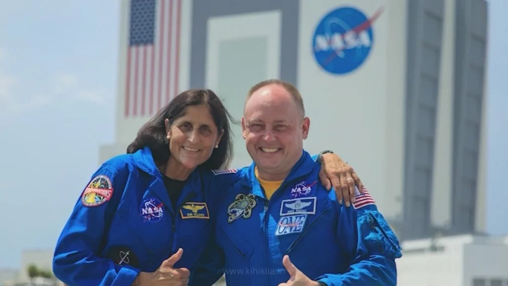 Sunita Williams and Butch Wilmore in white spacesuits shaking hands with NASA officials, standing near a rocket ahead of their space mission.